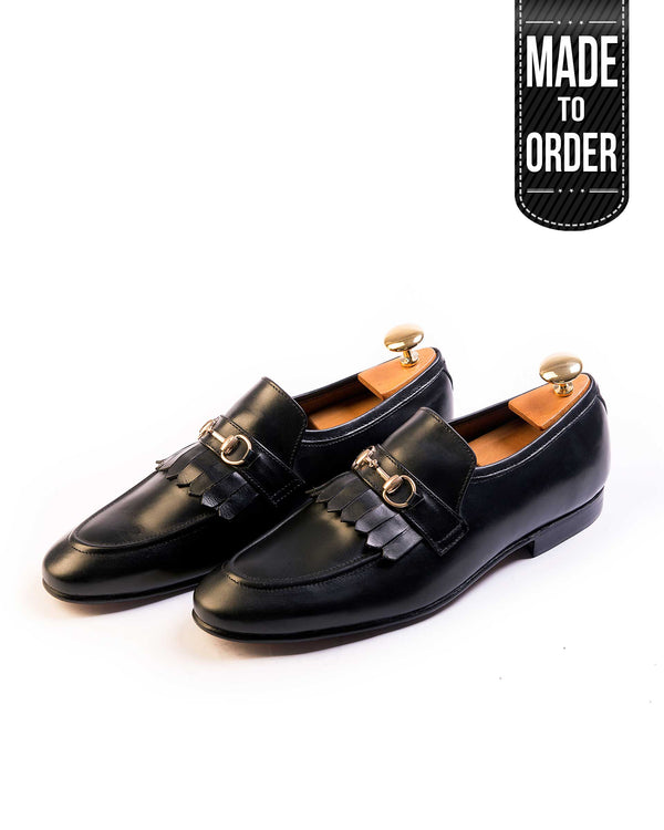 A pair of black leather moccasin shoes with a metal buckle on the top, displayed against a white background with a 'Made to Order' label on the top right.